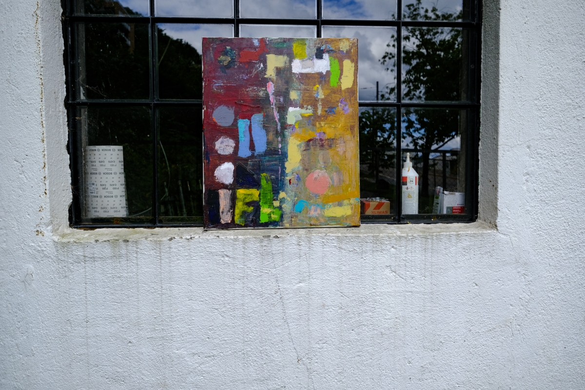 Abstract painting with red, yellow, blue, and green shapes resting on a window ledge, framed by a white textured wall.