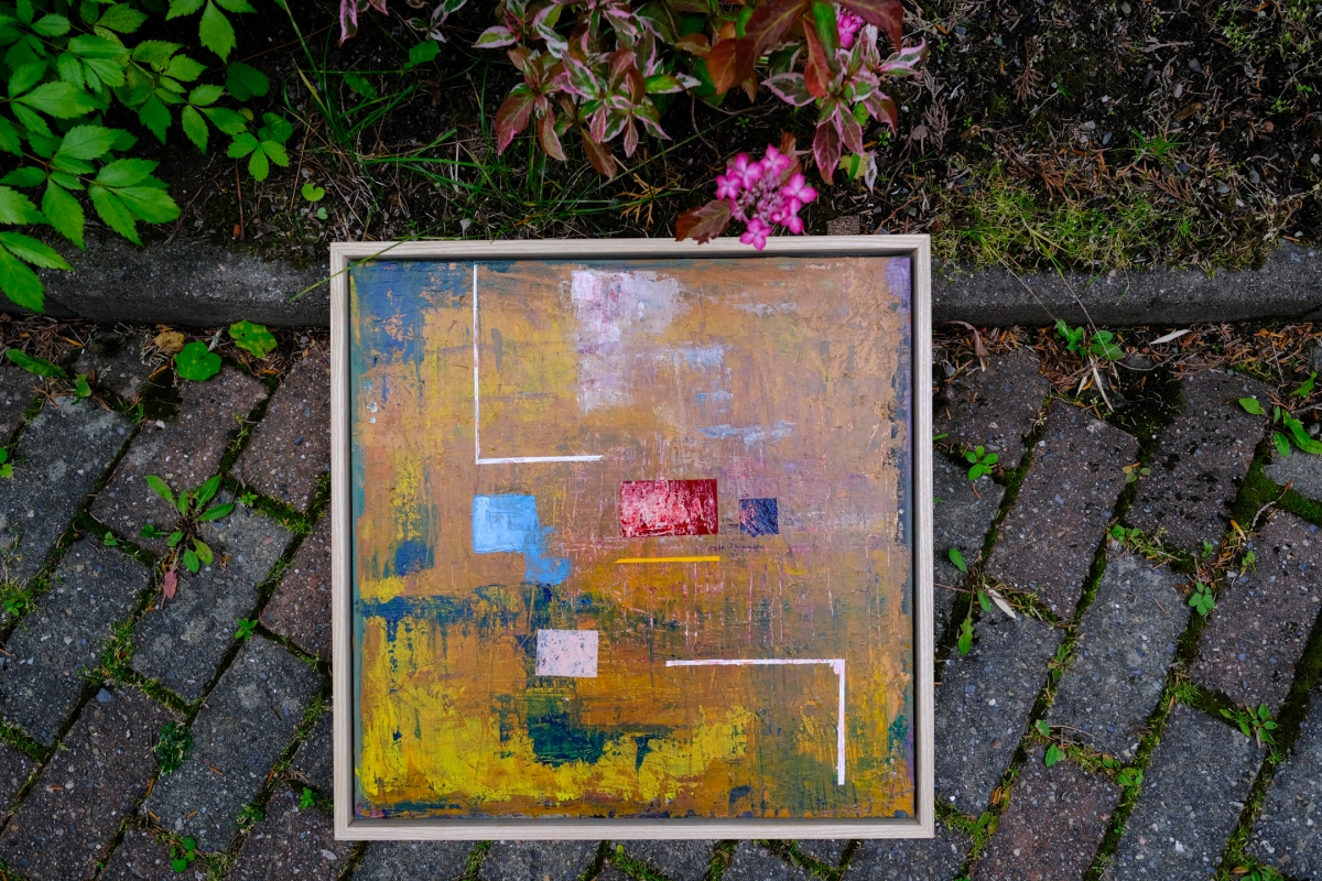 Abstract framed painting in ochre, gold, blue, and red tones with geometric shapes, resting on a stone path near green foliage and pink flowers.