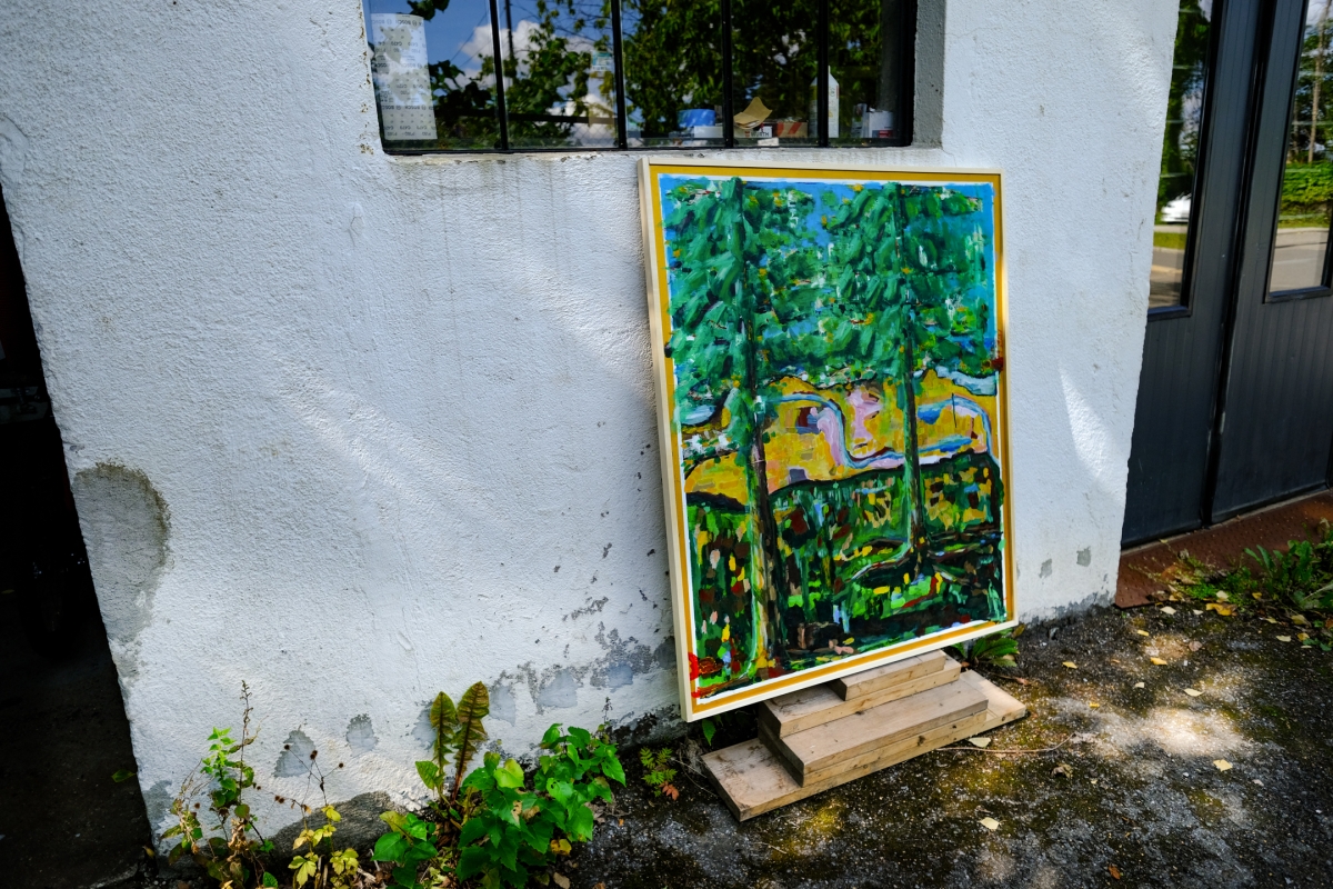 Colorful expressionist painting of tall pine trees against blue sky and yellow field, leaning on wooden blocks against a white stucco wall.