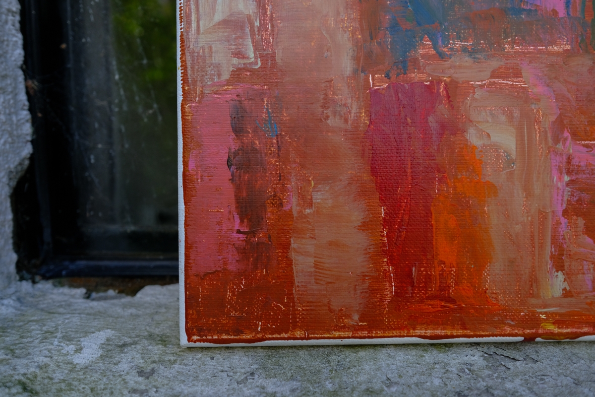 Abstract painting with red, pink, and orange brushstrokes resting on a weathered stone windowsill beside a dark-framed window.