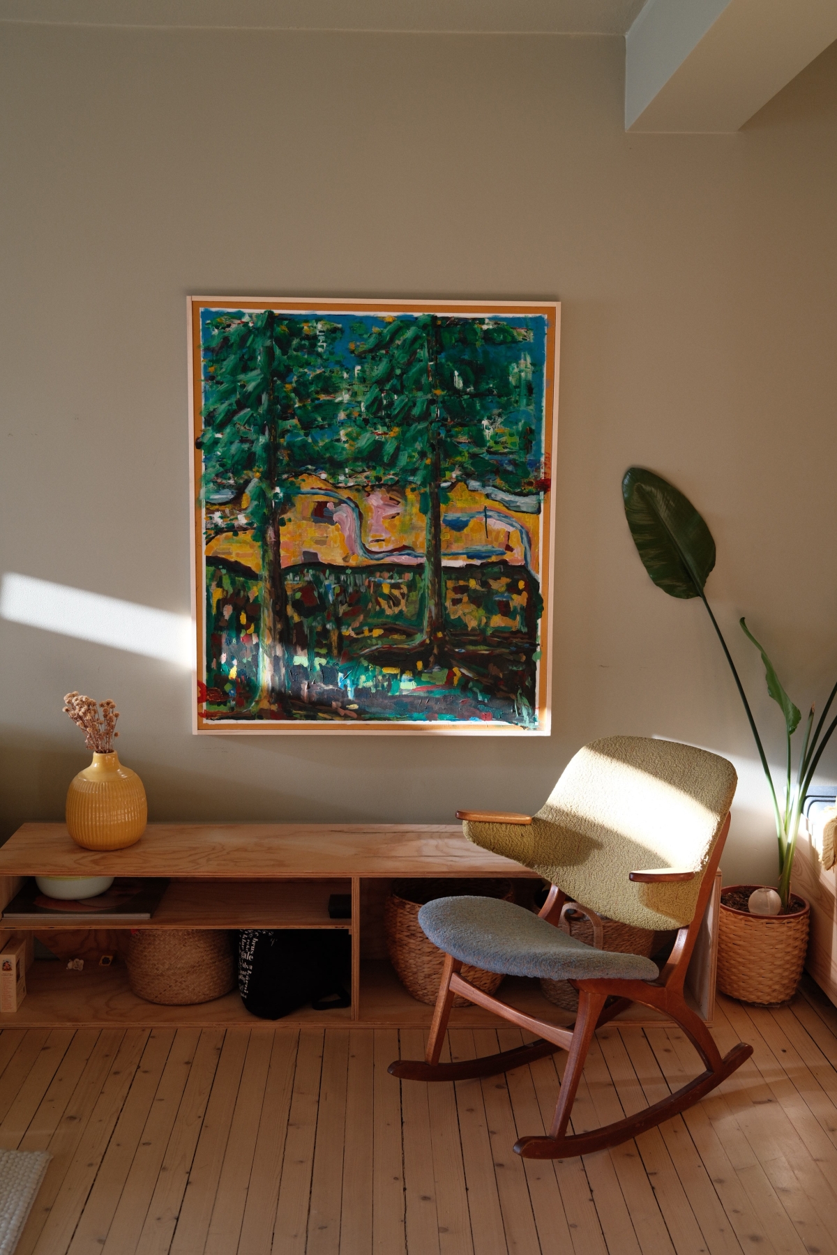 Sunlit living room with mid-century rocking chair, colorful tree painting, wooden console, yellow vase, and potted plant.