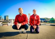Two men on top of a roof with red jackets and blue skies. 